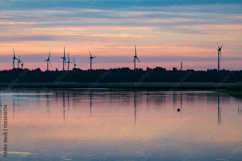 Evening sunlight on coast, pink and golden clouds and wind turbine. Sky reflection on water.  Wind generator for electricity, alternative energy source. Windmill for electric power production.