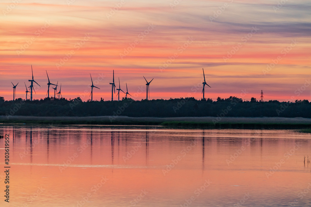 Evening sunlight on coast, pink and golden clouds and wind turbine. Sky reflection on water.  Wind generator for electricity, alternative energy source. Windmill for electric power production.