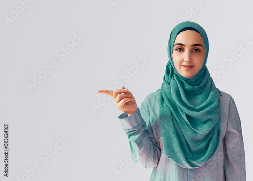 Cute Muslim woman with cheerful face expression points away with her forefinger, showing to copy space on the corner. Portrait of Muslim prayer woman in hijab