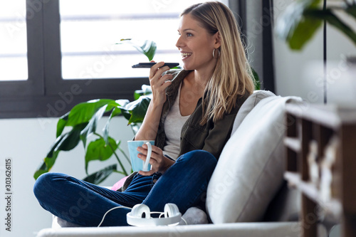 Pretty young woman talking on mobile phone with hands free while drinking cup of coffee on sofa at home. photo