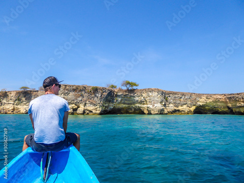 A man sitting on the prow of a blue boat and enjoying the idyllic view in front of him. There is a cliff formation emerging from the calm sea. The water has many shades of turquoise. Happiness