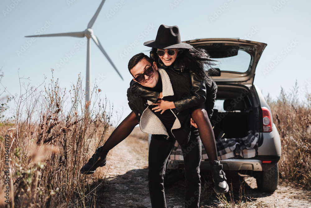 Obraz premium Happy Traveling Couple Enjoying a Car Trip on the Field Road with Electric Wind Turbine Power Generator on the Background
