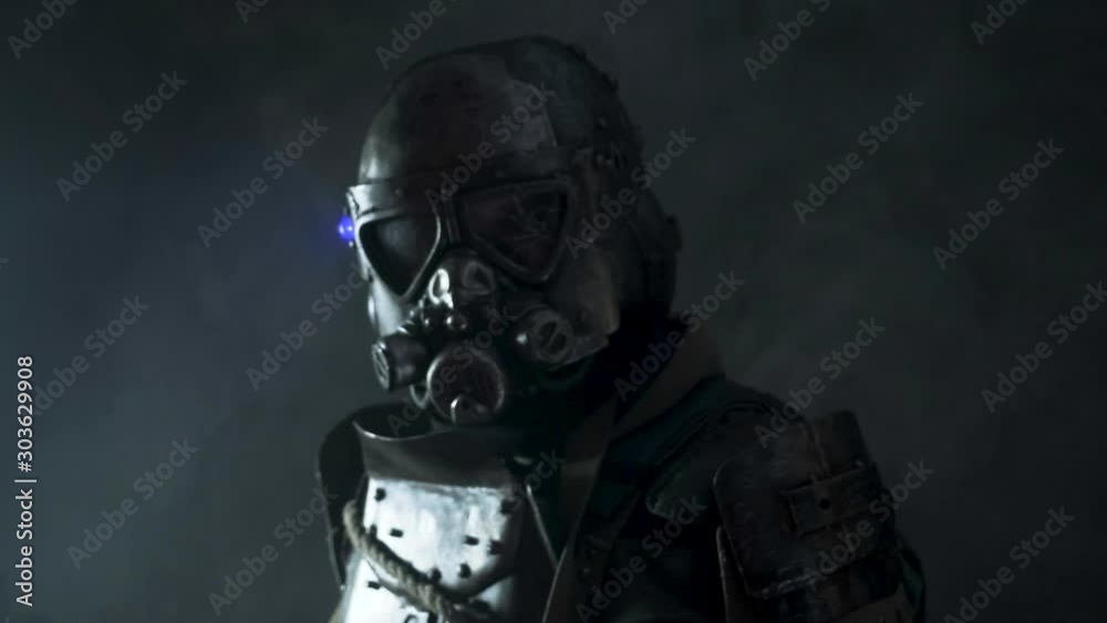 Apocalyptic warrior with weapon in full gear protective costume is standing at dark room. There are red flashlights reflecting on his gear. Apocalyptic warrior with weapon in full gear protective costume is standing at dark room. There are red flashlights reflecting on his gear.