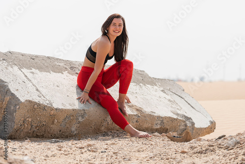 Beautiful brunette fitness model wearing a bright red tights and black sports bra sitting on a large slap of concrete in hot desert area on a bright day