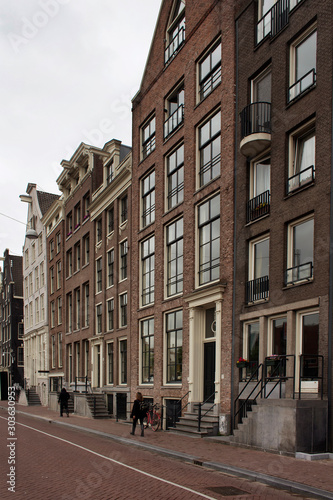 View of people walking in front of historical, traditional and typical buildings showing Dutch's architectural style in Amsterdam. It is a summer day with cloudy sky.