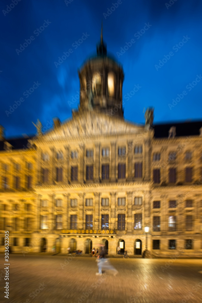 Blurry motion image of man walking on Dam square in Amsterdam. Royal Palace is in the background. It is a rainy summer night with cloudy, dark blue color sky.