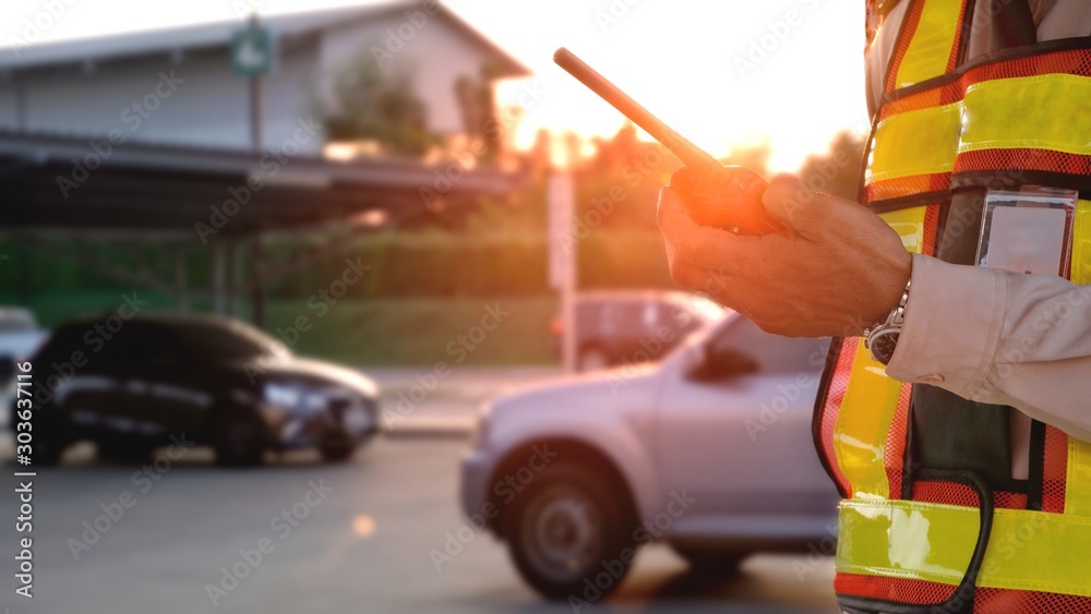 Security guard's hand in safety vest using walkie talkie or portable radio transmitter in outdoor parking lot area with flare light in evening time