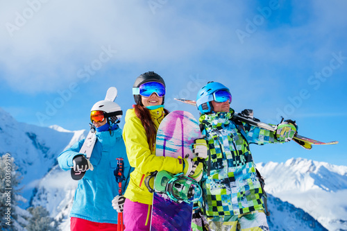 Happy men and woman with snowboard and skis looking in different directions standing on snow resort against background of mountain and cloudy sky.