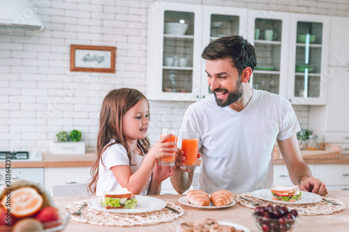 smiling father and daughter clicking glasses with drinking juice for breakfast in the kitchen