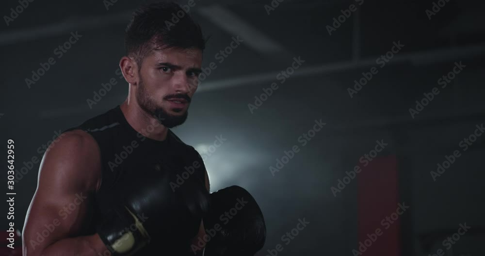 In a dark gym class brutal sweating guy with a big biceps doing his hard workout boxing exercises looking straight to the camera In a dark gym class brutal sweating guy with a big biceps doing his hard workout boxing exercises looking straight to the camera