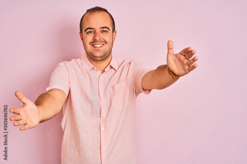 Young man wearing elegant shirt standing over isolated pink background looking at the camera smiling with open arms for hug. Cheerful expression embracing happiness. Young man wearing elegant shirt standing over isolated pink background looking at the camera smiling with open arms for hug. Cheerful expression embracing happiness.