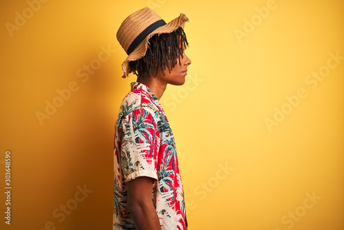 Afro american man with dreadlocks wearing floral shirt and hat over isolated yellow background looking to side, relax profile pose with natural face with confident smile.