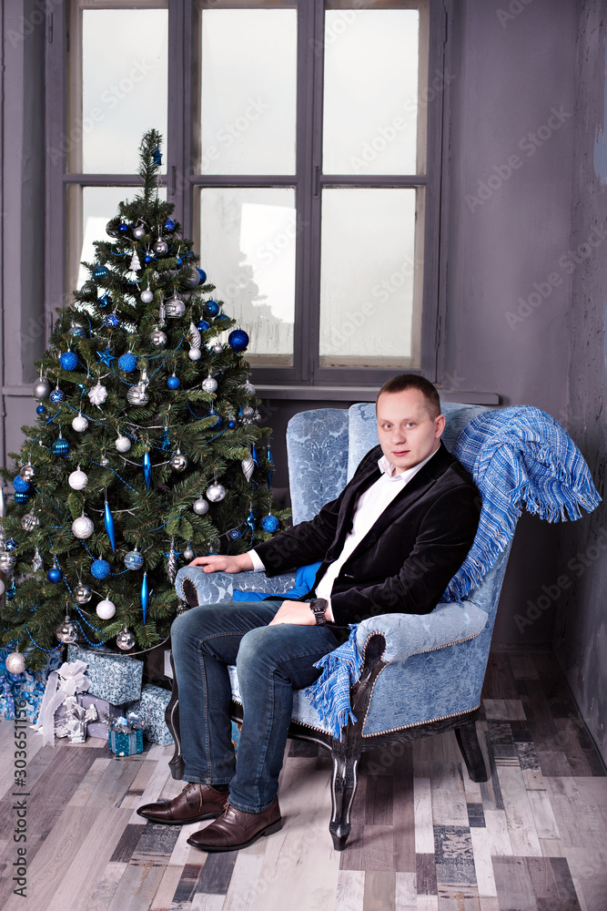 attractive young man sits on a chair near a decorated New year tree in Christmas