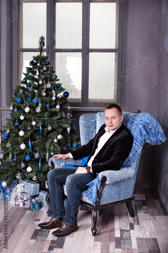 attractive young man sits on a chair near a decorated New year tree in Christmas © Tatsiana