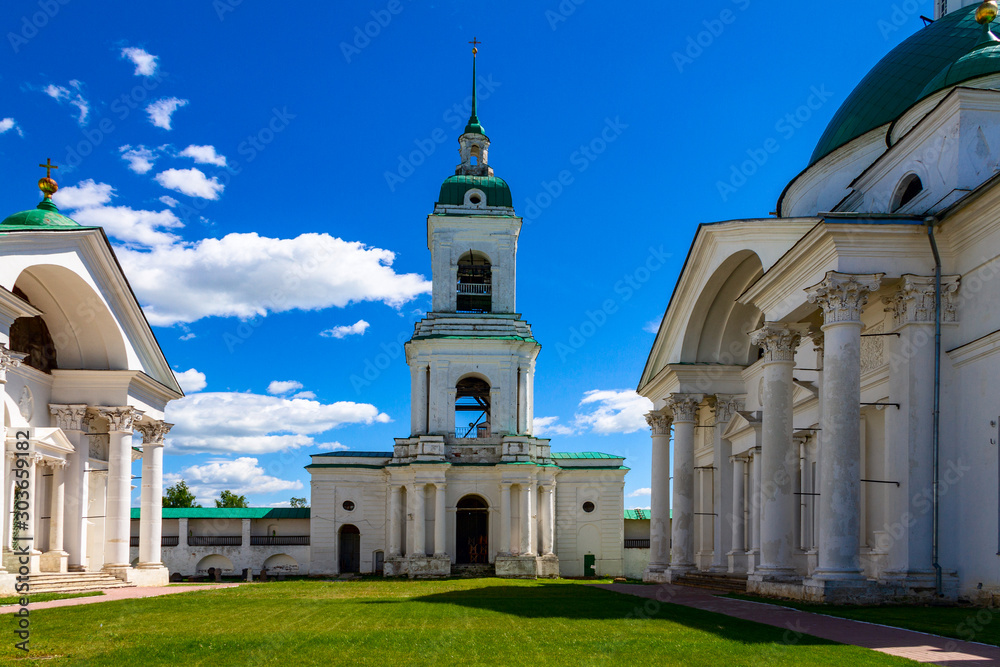 The bell tower of the Rostov Kremlin against the blue sky.  On the sides are the buildings of two churches.  All buildings are made of white stone.  Domes are green.