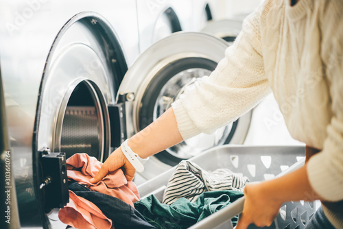 Interior of small laundromat in daylight. Close-up female holding basket. Girl loading dirty clothes inside drum and closing door. Self-service concept