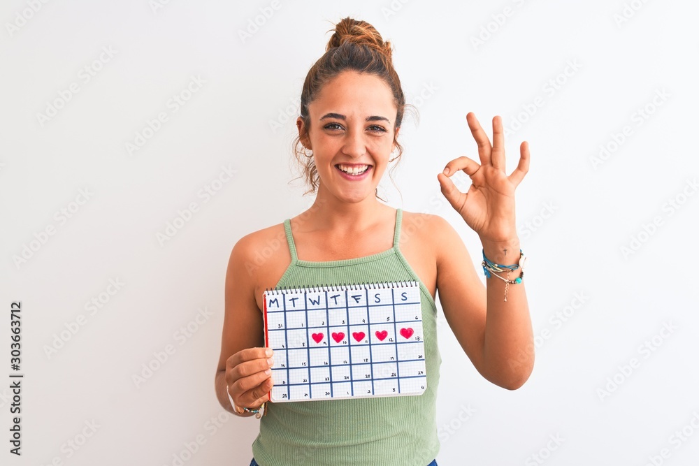 Young redhead woman holding menstruation calendar over isolated background doing ok sign with fingers, excellent symbol Young redhead woman holding menstruation calendar over isolated background doing ok sign with fingers, excellent symbol
