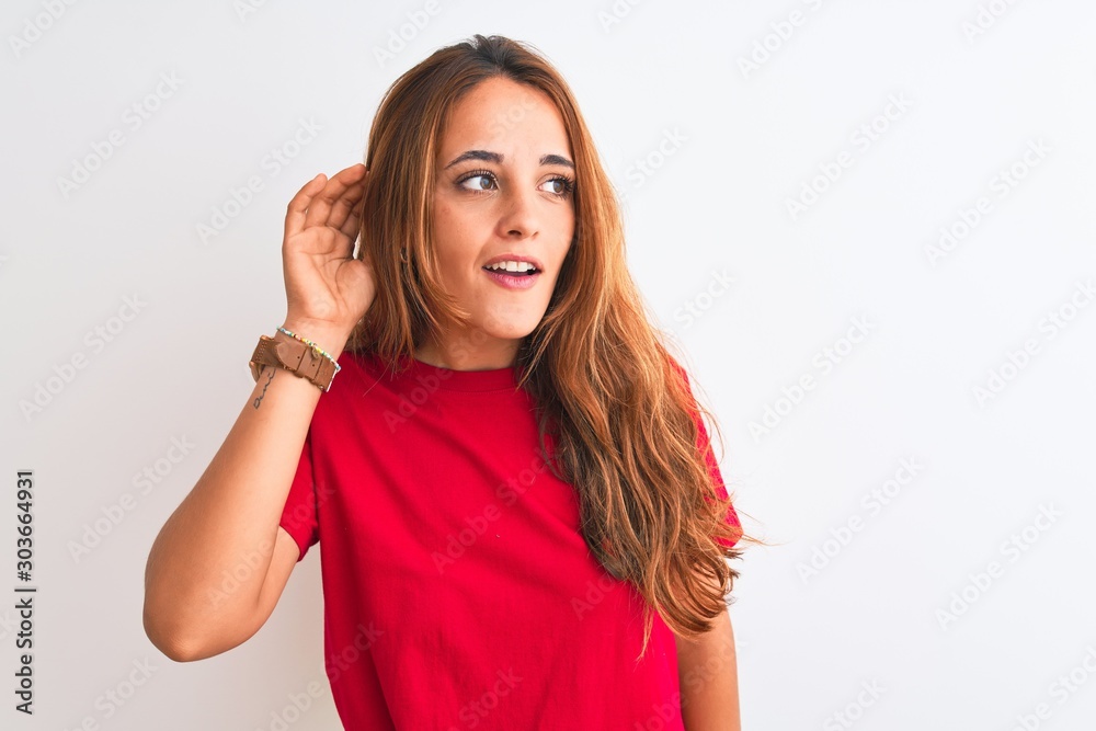 Young redhead woman wearing red casual t-shirt stading over white isolated background smiling with hand over ear listening an hearing to rumor or gossip. Deafness concept. Young redhead woman wearing red casual t-shirt stading over white isolated background smiling with hand over ear listening an hearing to rumor or gossip. Deafness concept.