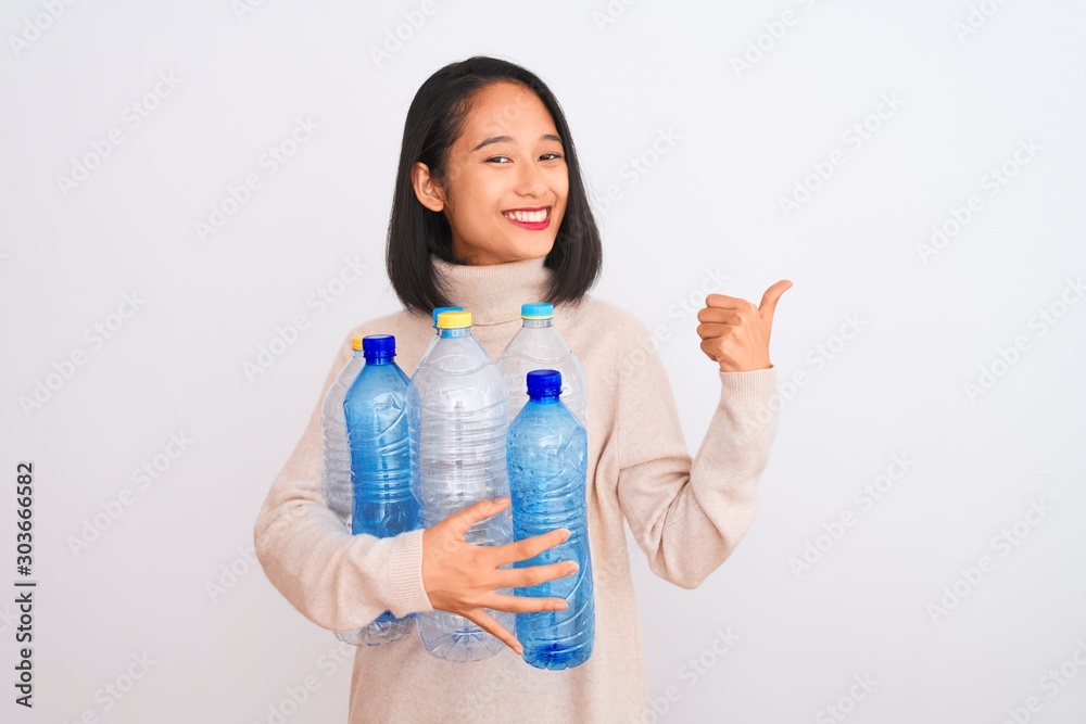 Young beautiful chinese woman recycling plastic bottles over isolated white background happy with big smile doing ok sign, thumb up with fingers, excellent sign Young beautiful chinese woman recycling plastic bottles over isolated white background happy with big smile doing ok sign, thumb up with fingers, excellent sign