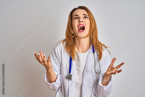 Redhead caucasian doctor woman wearing stethoscope over isolated background crazy and mad shouting and yelling with aggressive expression and arms raised. Frustration concept.