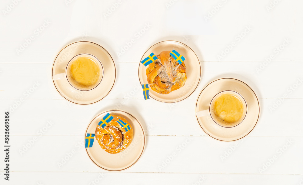 Swedish cinnamon buns (kanelbullar) and two cups of coffee on the white wooden table. Swedish flags. Coffee break (fika) concept. Top view. Swedish cinnamon buns (kanelbullar) and two cups of coffee on the white wooden table. Swedish flags. Coffee break (fika) concept. Top view.