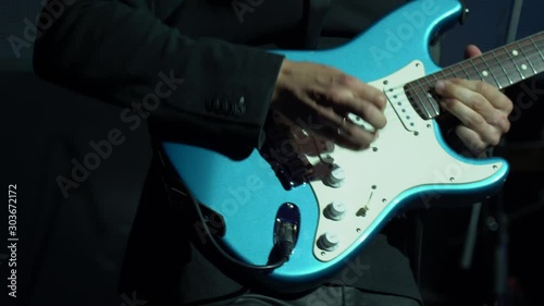 Handsome white man guitar player in a black jacket quickly and actively play with pleasure on a blue and white professional electric guitar on a black background in the light of spotlights close up.