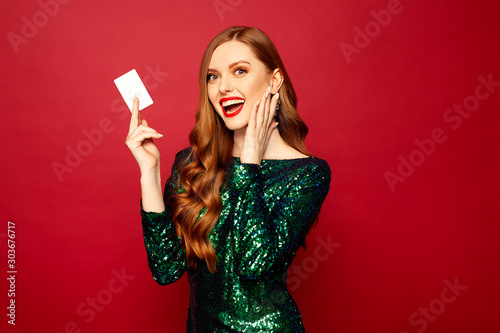 beautiful red-haired model stands on a red background and smiles a beautiful smile in brilliant dress, a card in hands , merry christmas and happy new year