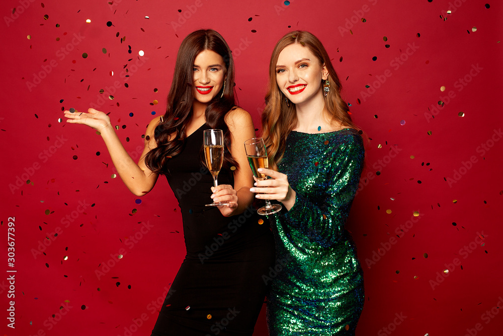 Plakat dwie piękne modelki, ruda i brunetka w noworocznych sukienkach, bawiących się i uśmiechających się z kieliszkami szampana, konfetti latającymi po czerwonym tle. Nowy rok lub zdjęcie świąteczne