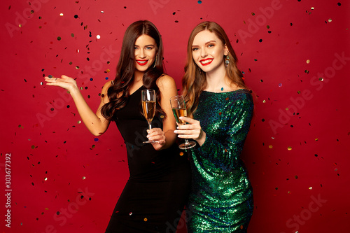  two beautiful models, a redhead and a brunette in New Year's dresses, having fun and smiling with glasses of champagne, confetti flying around on a red background. New Year or Christmas photo