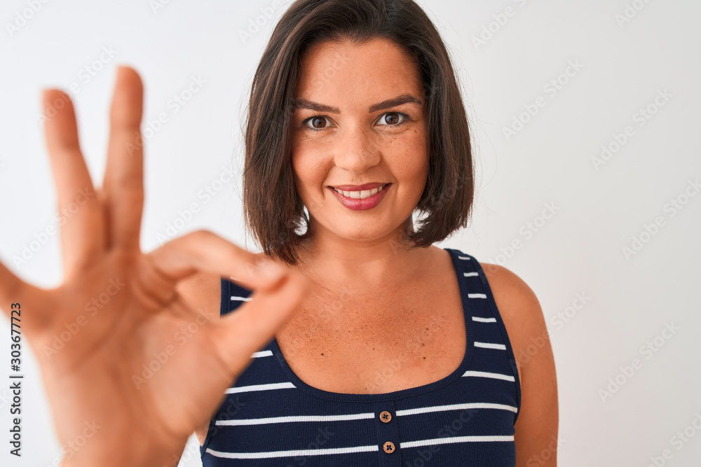 Young beautiful woman wearing blue striped t-shirt standing over isolated white background doing ok sign with fingers, excellent symbol Young beautiful woman wearing blue striped t-shirt standing over isolated white background doing ok sign with fingers, excellent symbol