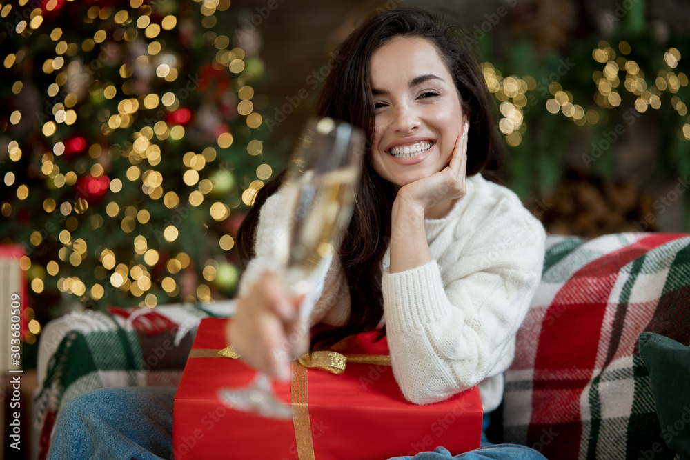Young smiling woman wearing cozy sweater holding present and champagne sitting on the sofa in room decorated for celebrating new year and christmas festive mood Young smiling woman wearing cozy sweater holding present and champagne sitting on the sofa in room decorated for celebrating new year and christmas festive mood