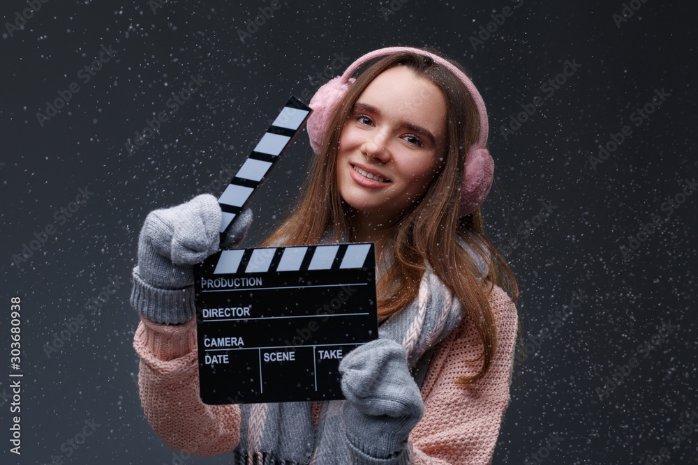 Young beautiful smiling girl in knitting pullover holding director's clapper board for making Christmas video in studio.Movie production clapper board.Lights, camera, action. Start of winter holidays.