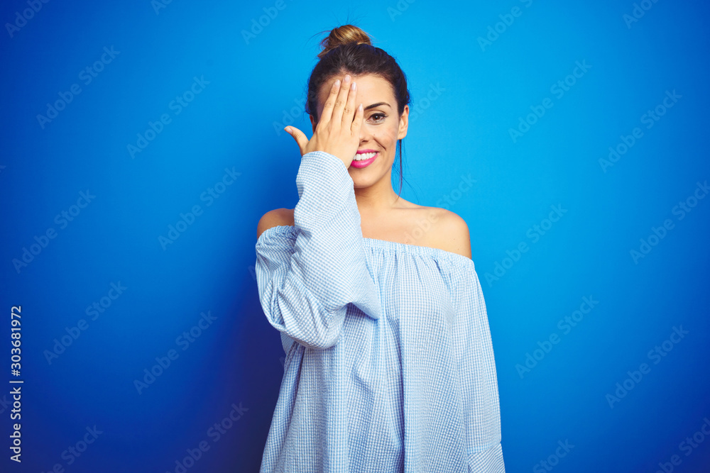 Young beautiful woman wearing bun hairstyle over blue isolated background covering one eye with hand, confident smile on face and surprise emotion. Young beautiful woman wearing bun hairstyle over blue isolated background covering one eye with hand, confident smile on face and surprise emotion.