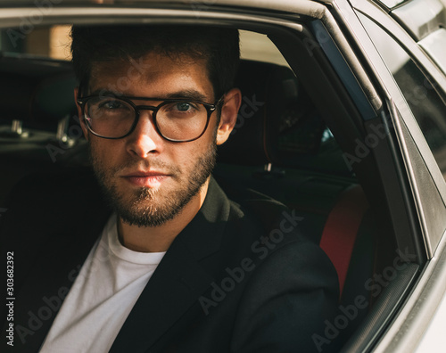 Attractive young man with beard and glasses with serious face looks at camera through the window of his car. Businessman. © Freestocker