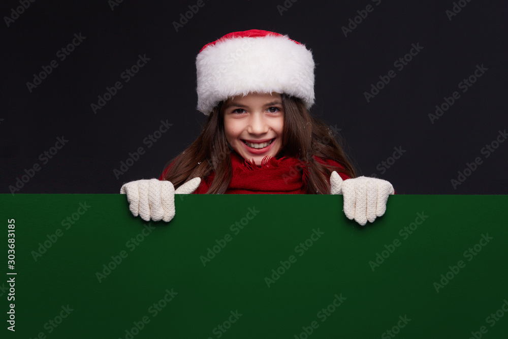 Young beautiful smiling girl in red Santa hat holding empty green banner with copy space. Kid hiding behind card blank. Winter holidays, Christmas, New Year concept. Young beautiful smiling girl in red Santa hat holding empty green banner with copy space. Kid hiding behind card blank. Winter holidays, Christmas, New Year concept.