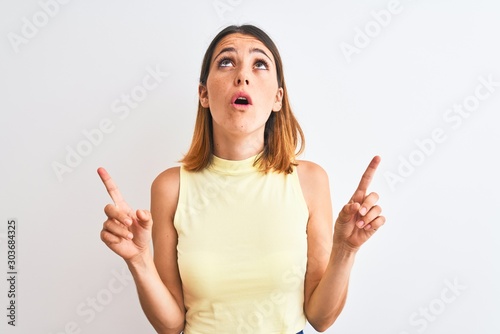Beautiful redhead woman wearing yellow summer t-shirt over isolated background amazed and surprised looking up and pointing with fingers and raised arms.