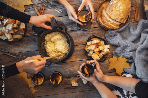 Top view friends cooking Swiss fondue assorted cheeses dinner on fire, hands holding mulled wine background wood board