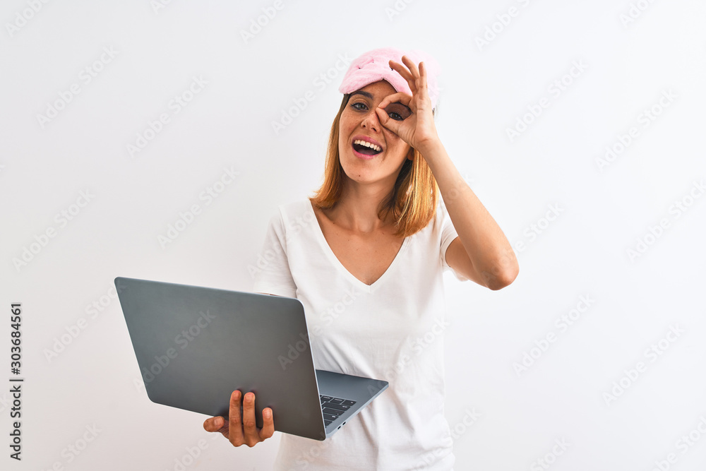 Beautiful redhead woman wearing sleeping mask using laptop over isolated background with happy face smiling doing ok sign with hand on eye looking through fingers Beautiful redhead woman wearing sleeping mask using laptop over isolated background with happy face smiling doing ok sign with hand on eye looking through fingers