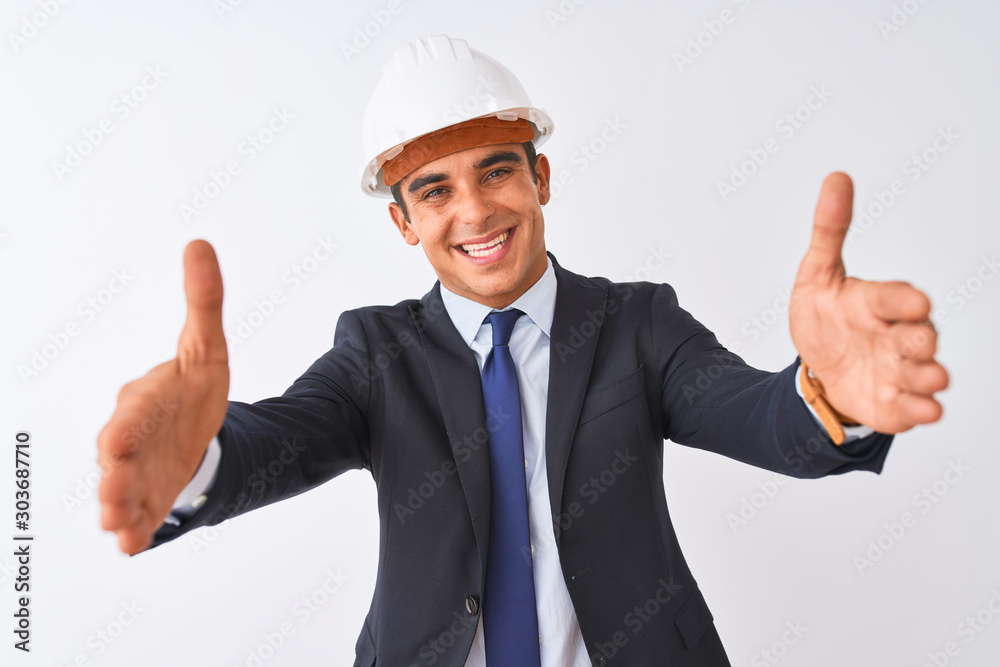 Young handsome architect man wearing suit and helmet over isolated white background looking at the camera smiling with open arms for hug. Cheerful expression embracing happiness.