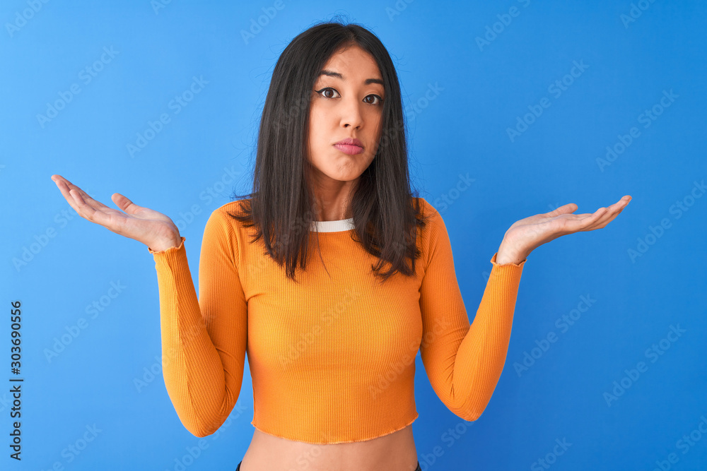 Φωτογραφία Young beautiful chinese woman wearing orange t-shirt standing over isolated blue background clueless and confused expression with arms and hands raised