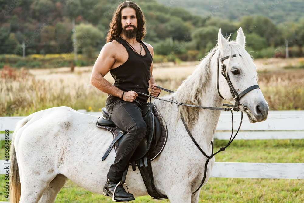 Obraz premium Young jockey training his horse for a ride. Good looking, hunky cowboy rides horse at beautiful country side nature. Equestrian rides his white well looking horse.