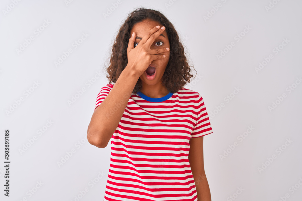 Young brazilian woman wearing red striped t-shirt standing over isolated white background peeking in shock covering face and eyes with hand, looking through fingers with embarrassed expression. Young brazilian woman wearing red striped t-shirt standing over isolated white background peeking in shock covering face and eyes with hand, looking through fingers with embarrassed expression.