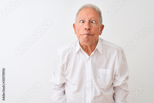 Senior grey-haired man wearing elegant shirt standing over isolated white background making fish face with lips, crazy and comical gesture. Funny expression.
