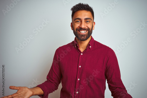 Young indian man wearing red elegant shirt standing over isolated grey background smiling cheerful with open arms as friendly welcome, positive and confident greetings
