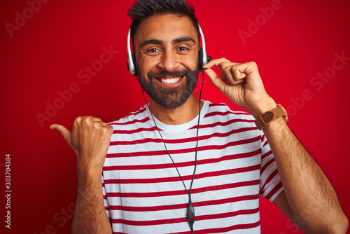 Young indian call center agent man using headset over isolated red background pointing and showing with thumb up to the side with happy face smiling