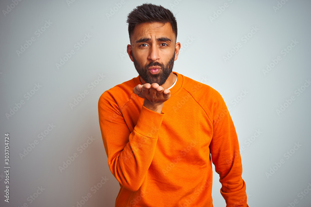 Young indian man wearing orange sweater over isolated white background looking at the camera blowing a kiss with hand on air being lovely and sexy. Love expression. Young indian man wearing orange sweater over isolated white background looking at the camera blowing a kiss with hand on air being lovely and sexy. Love expression.