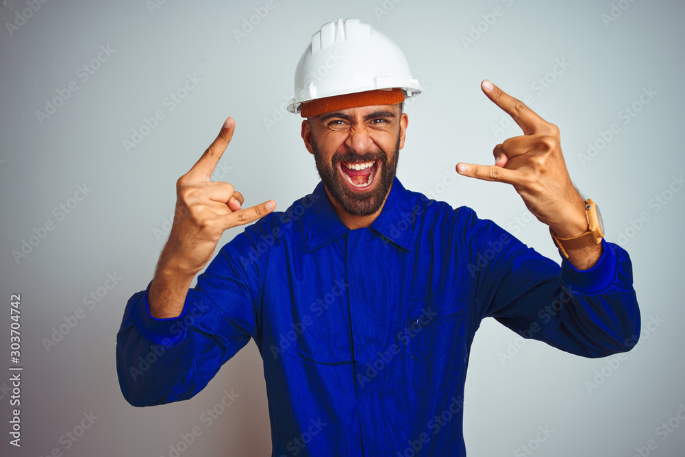 Handsome indian worker man wearing uniform and helmet over isolated white background shouting with crazy expression doing rock symbol with hands up. Music star. Heavy concept.