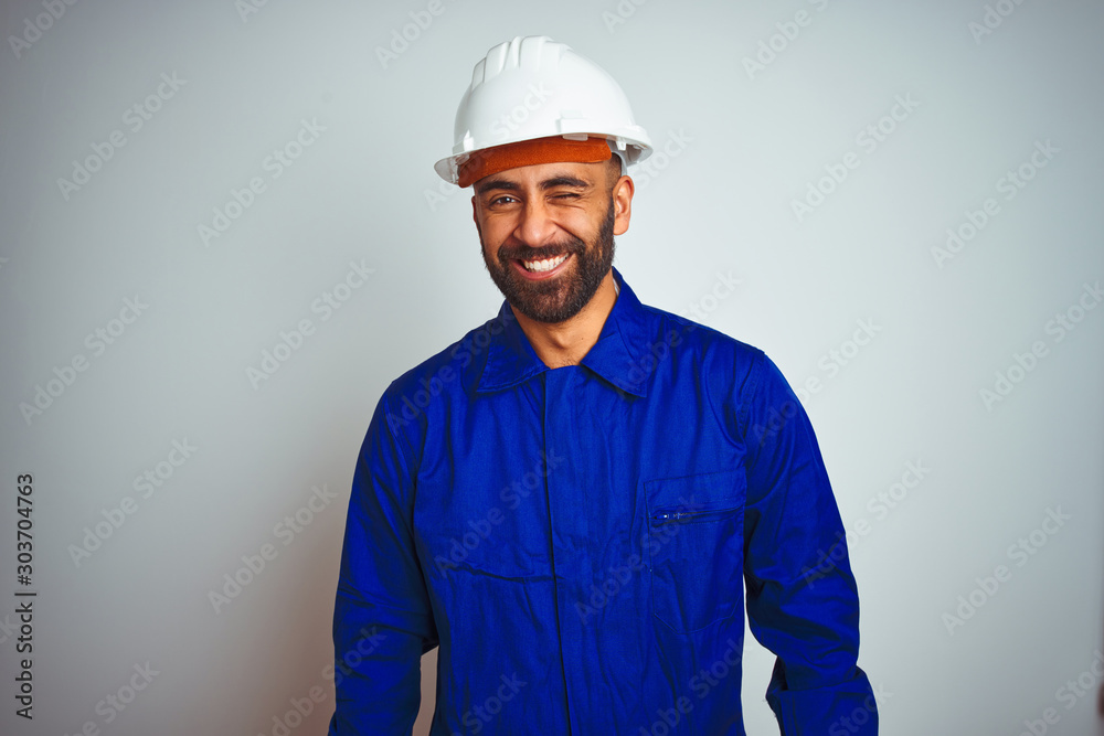 Handsome indian worker man wearing uniform and helmet over isolated white background winking looking at the camera with sexy expression, cheerful and happy face.