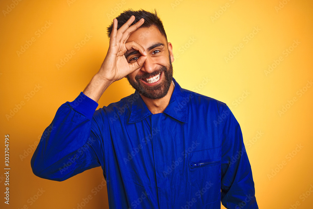 Handsome indian worker man wearing uniform over isolated yellow background doing ok gesture with hand smiling, eye looking through fingers with happy face. Handsome indian worker man wearing uniform over isolated yellow background doing ok gesture with hand smiling, eye looking through fingers with happy face.