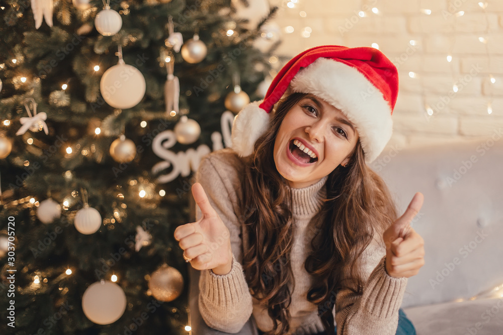 Woman in santa hat showing thumbs up sitting on the sofa near Christmas tree in the decorative interior. Positive gesture. Christmas and New Year photo.